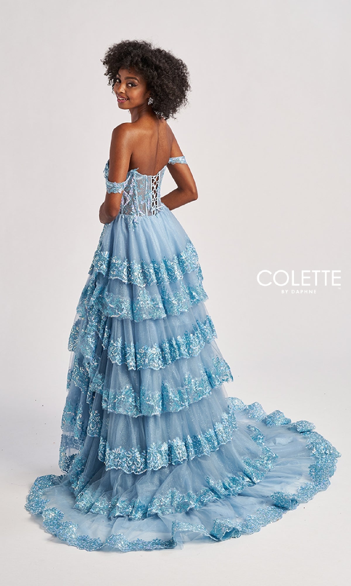 A woman in the Colette CL8690 long prom dress, blue and off-shoulder with lace details, stands partly turned away. The gown has a corset back, ruffle-tiered A-line skirt, and a dramatic flowing train.